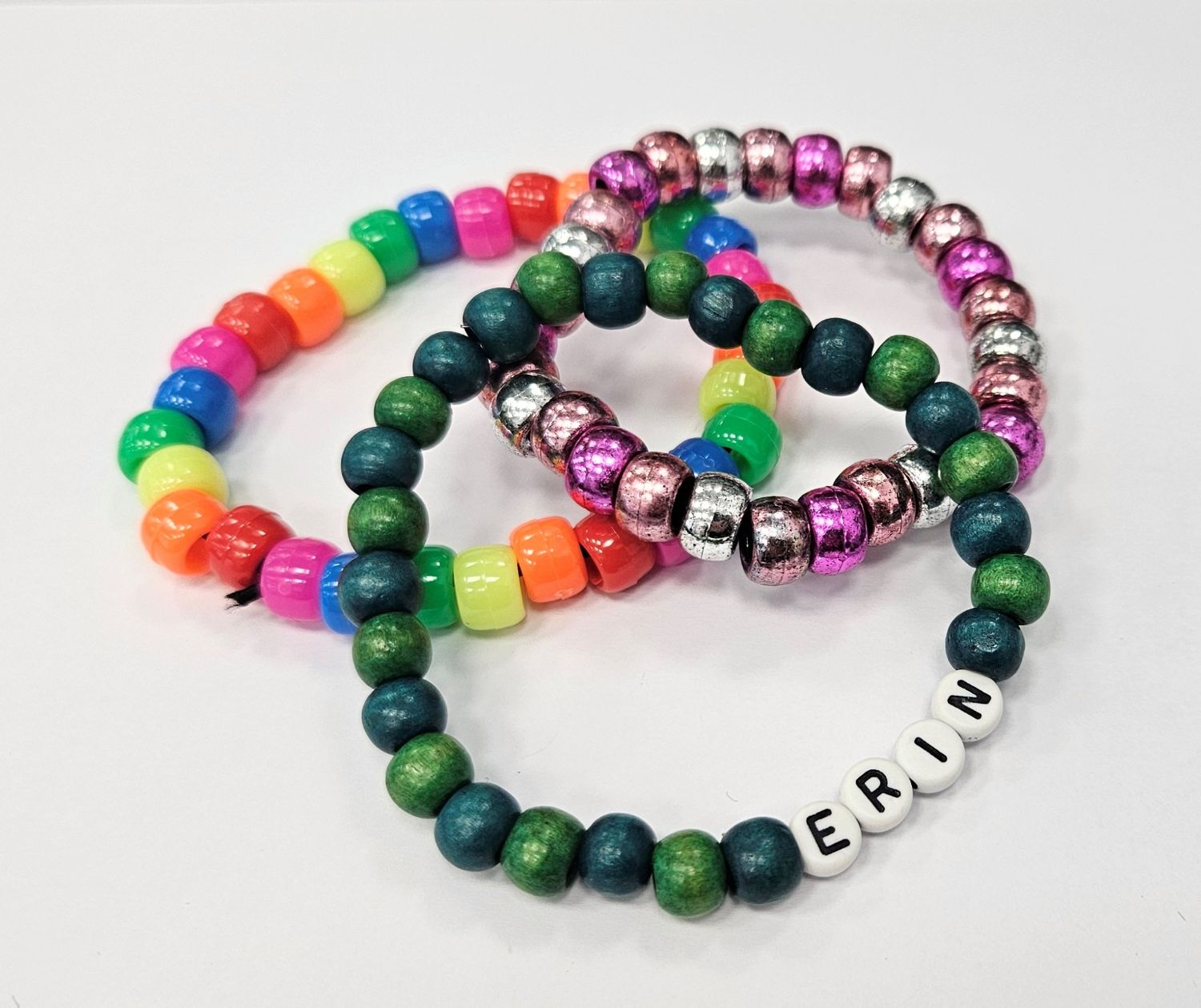 Three distinct handmade beaded bracelets are grouped on a white background, including a rainbow pony bead design, a shiny metallic pink and silver band, and a green wooden bead bracelet.